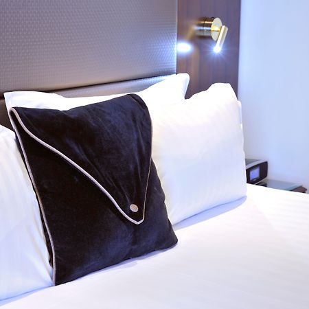 Stamford Plaza Melbourne Hotel Ngoại thất bức ảnh The photo shows a neatly arranged bed with white linens. There are several pillows, with one black pillow featuring a distinctive design on top of the white pillows. The headboard of the bed is visible, along with a stylish wall-mounted light fixture