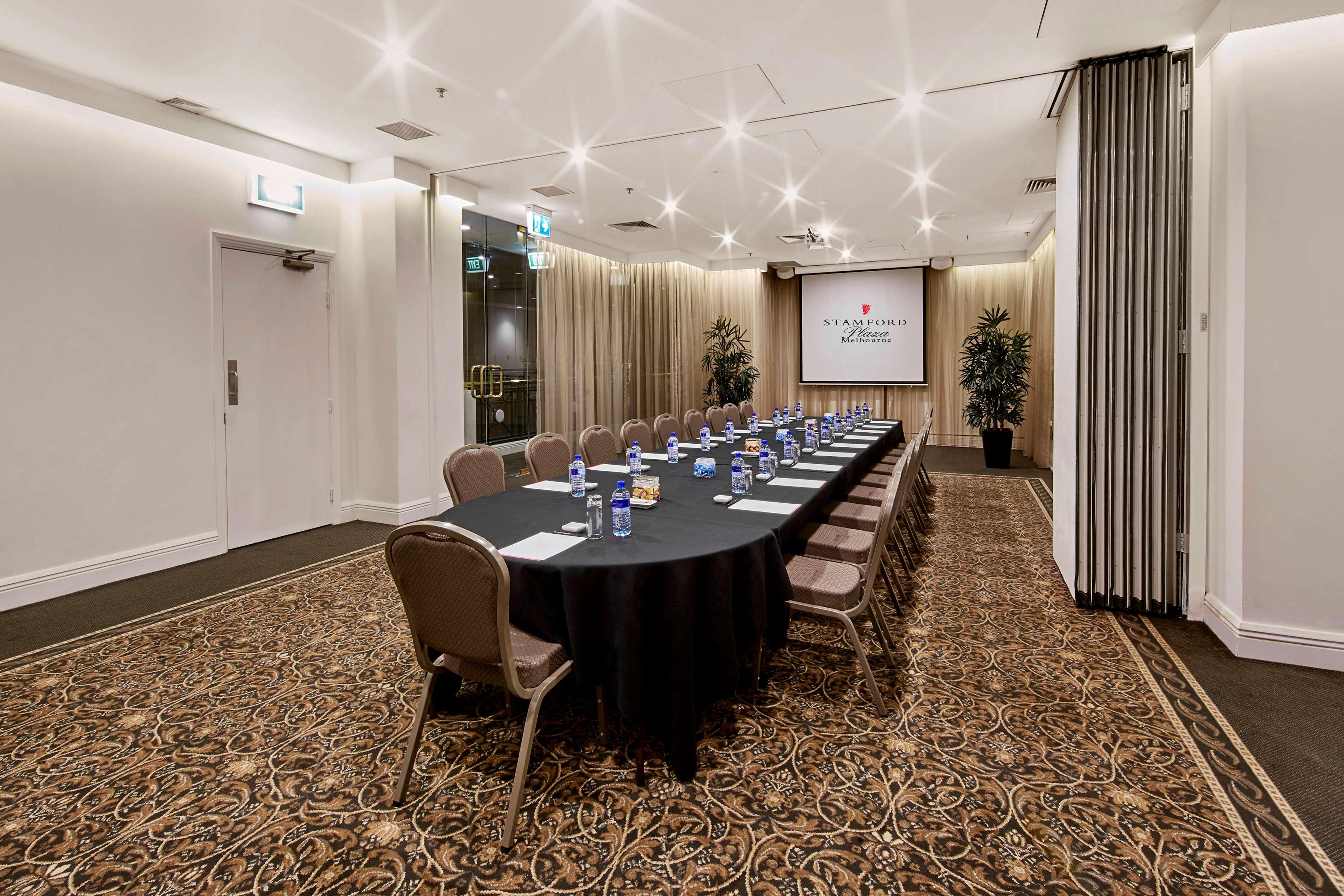 Stamford Plaza Melbourne Hotel Ngoại thất bức ảnh The photo shows a meeting room set up for a conference or presentation. There is a long, rectangular table covered with a black tablecloth, surrounded by chairs. Each seat has a water bottle placed in front of it. At the front of the room, there is a