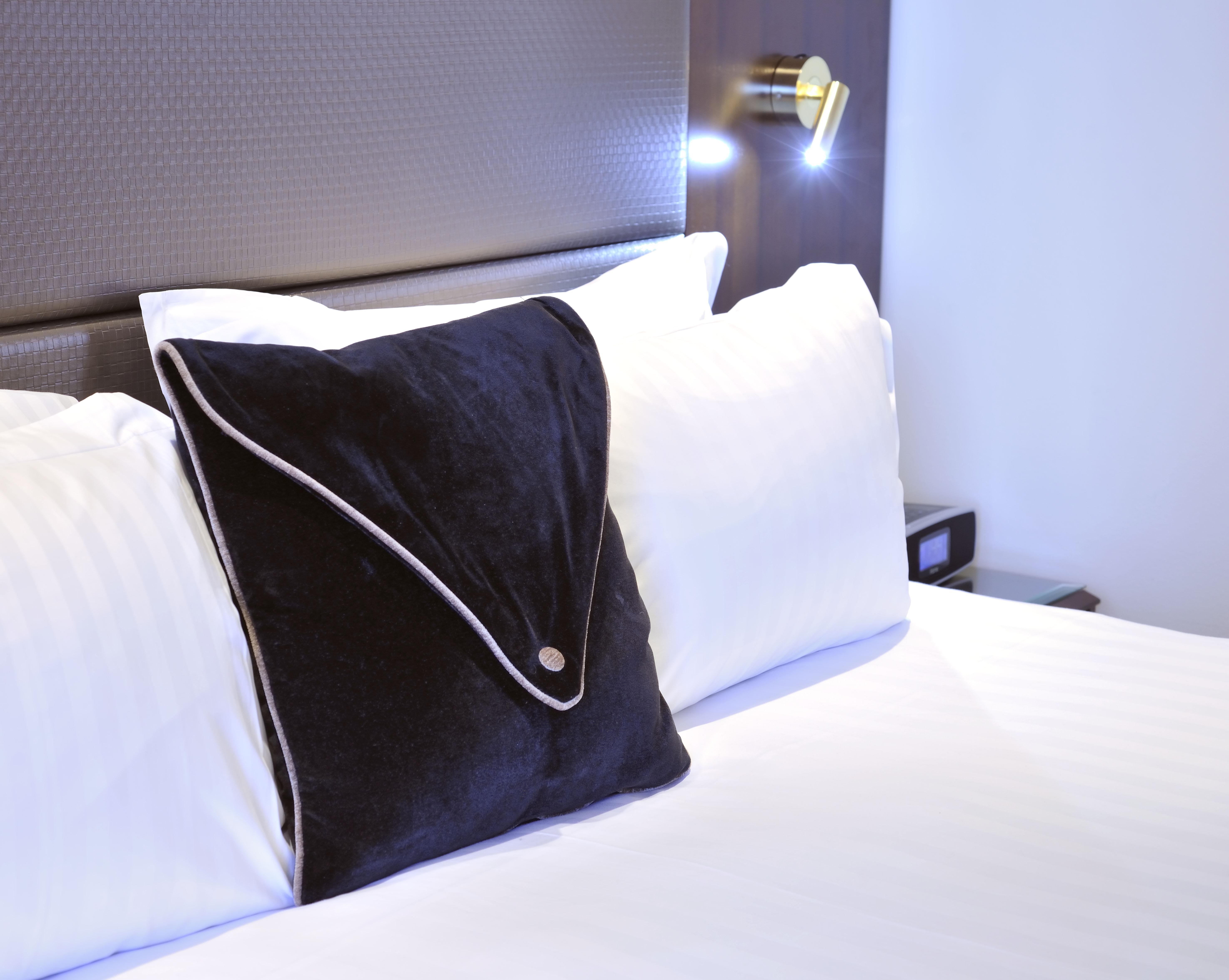 Stamford Plaza Melbourne Hotel Ngoại thất bức ảnh The photo shows a neatly arranged bed with white linens. There are several pillows, with one black pillow featuring a distinctive design on top of the white pillows. The headboard of the bed is visible, along with a stylish wall-mounted light fixture