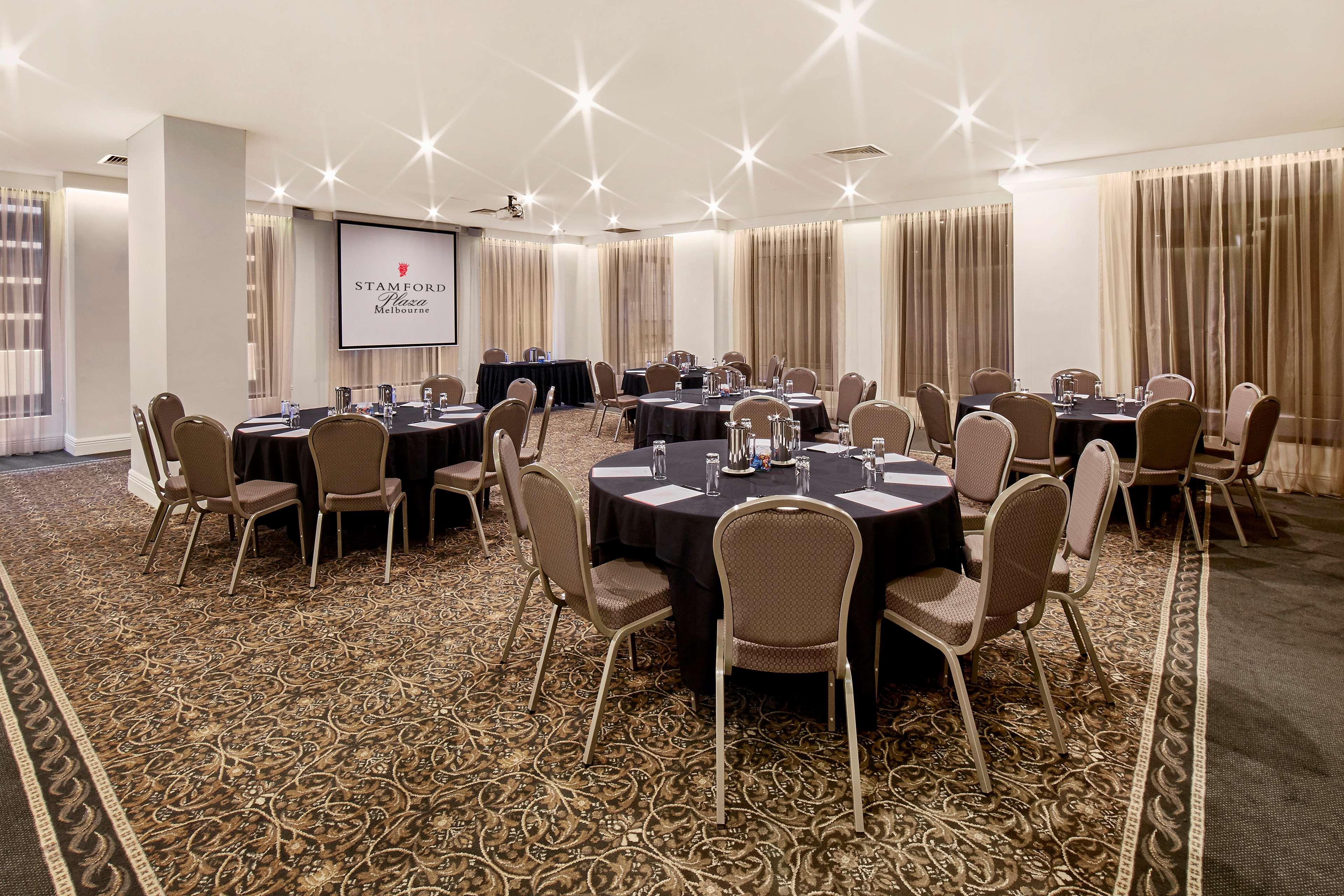 Stamford Plaza Melbourne Hotel Ngoại thất bức ảnh The photo shows a well-lit event space set up for a gathering or meeting. There are several round tables covered with black tablecloths, each surrounded by beige chairs. In the background, a large screen displays a logo or branding, likely related to