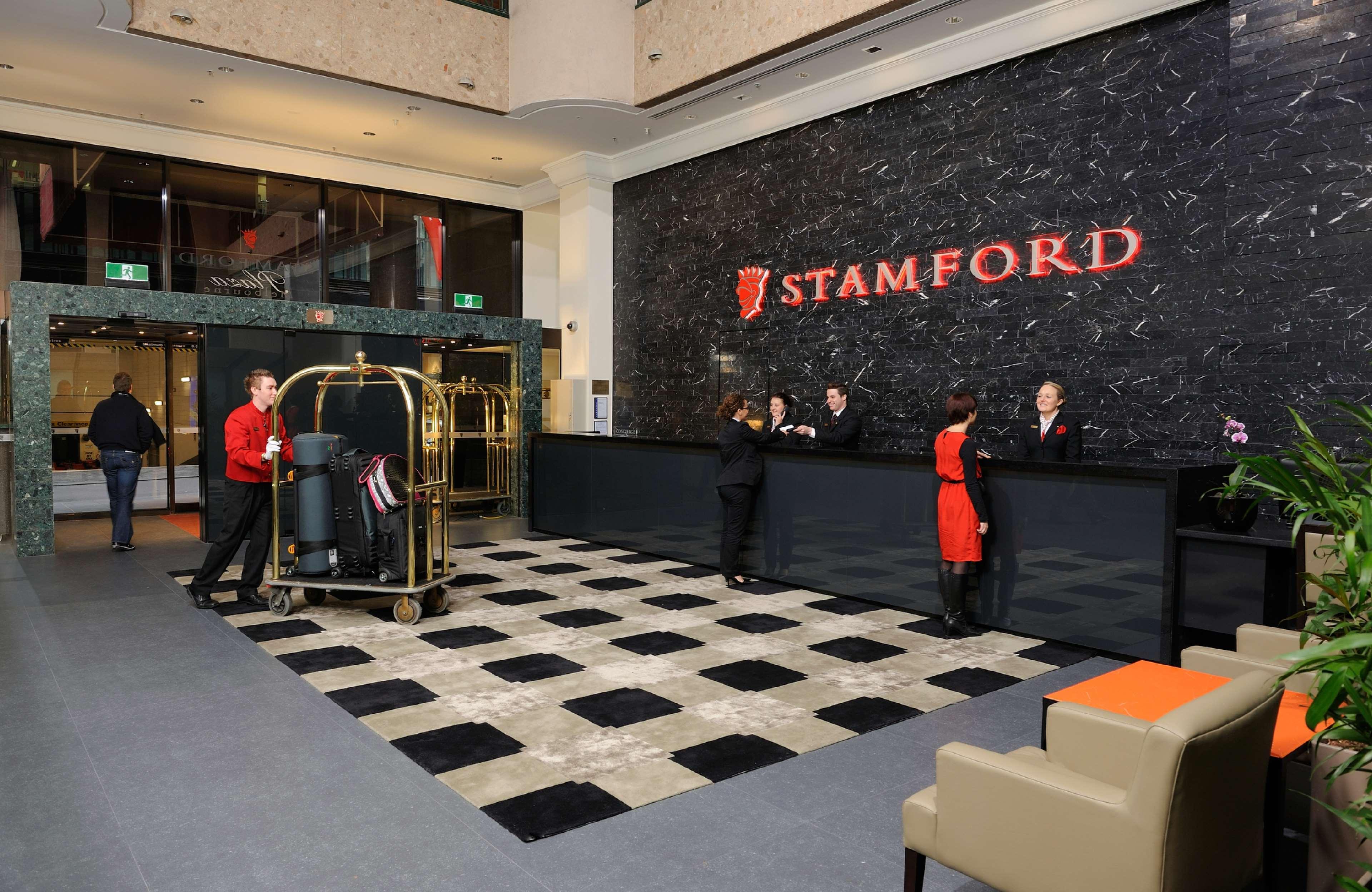 Stamford Plaza Melbourne Hotel Nội địa bức ảnh The photo shows the interior of a hotel lobby, specifically the Stamford Hotel. There is a large reception desk where several staff members are attending to guests. In the foreground, a bell cart loaded with luggage is positioned beside the reception