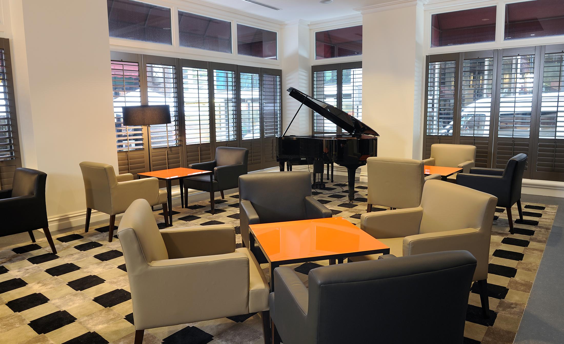 Stamford Plaza Melbourne Hotel Ngoại thất bức ảnh The photo shows a stylish indoor lounge area. It features several light gray armchairs arranged around orange square tables. In the corner, there is a black grand piano, adding an elegant touch to the space. The room has large windows with wooden shu
