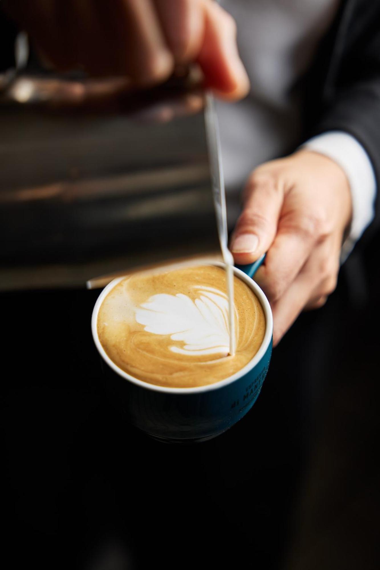 Stamford Plaza Melbourne Hotel Ngoại thất bức ảnh The photo shows a close-up of a hand holding a cup of coffee with a latte art design on the surface. The coffee appears to be a creamy, caramel color, likely a latte or cappuccino. A small stream of milk is being poured into the cup, creating a heart
